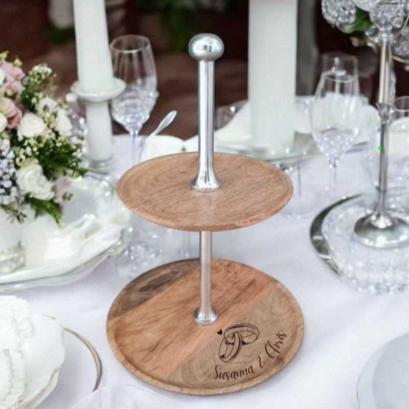 Moderne zweistöckige Etagere aus rustikalem Holz mit Stab und Knopfgriff aus silbernem Aluminium, dekoriert auf einem Hochzeitstisch. Ein Motiv zum Beispiel mit Herz und Ringen und frei wählbarer Wunschtext kann per Lasergravur hinzugefügt werden.