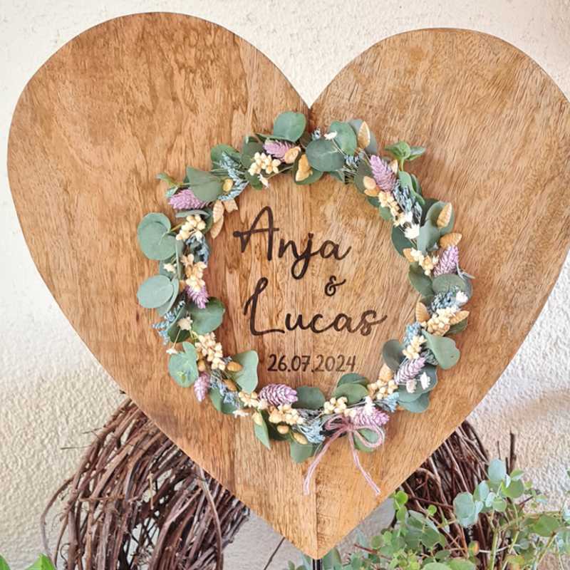 Detailansicht zur Gravur eines Holzherzens zur Hochzeit. Es ist ein großes Herz auf massivem Metallständer, mit Namen und Datum individuell auf Wunsch graviert. Ein Blumenkranz umrankt den Wunschtext. Toll als Deko oder Geschenk fürs Brautpaar.