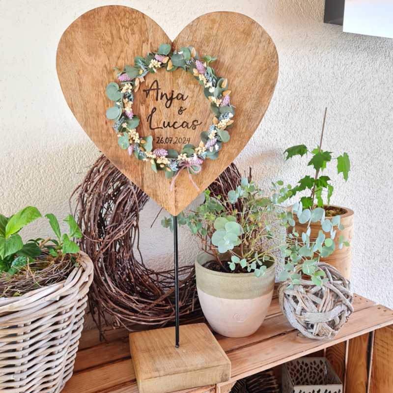 Ein großes Holzherz wird extra zur Hochzeit graviert. Der Wunschtext ist mittig aufgebracht und wird von einem Kranz aus Grün und Trockenblumen romantisch umrahmt. Ein individuelles und besonderes Geschenk zur Trauung.