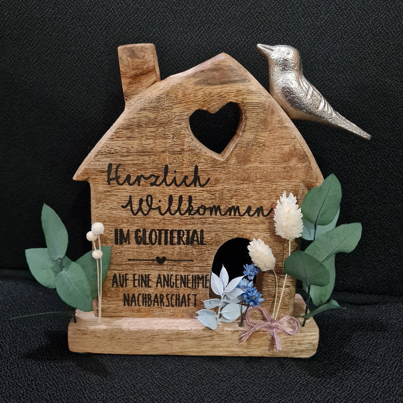 ein Geschenk zum Einzug, für neue Nachbarn oder zum Umzug. Ein kleines Haus aus Holz, mit Trockenblumen dekoriert und mit Wunschtext graviert. Auf gute Nachbarschaft. Ein kleiner Silbervogel sitzt auf dem Dach. Schlicht und schön.