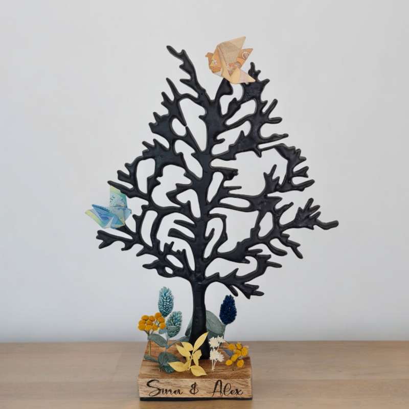Der Baum aus Metall steht auf einem gravierten Holzsockel. Dieser wird mit Namen und Datum oder kurzem Wunschtext individuell beschriftet. Die Geldscheine werden zu Tauben gefaltet und sitzen im Baum. Der Baum steht in einer gesteckten Wiese aus Trockenblumen. Eine wunderschöne Idee als Hochzeitsgeschenk.