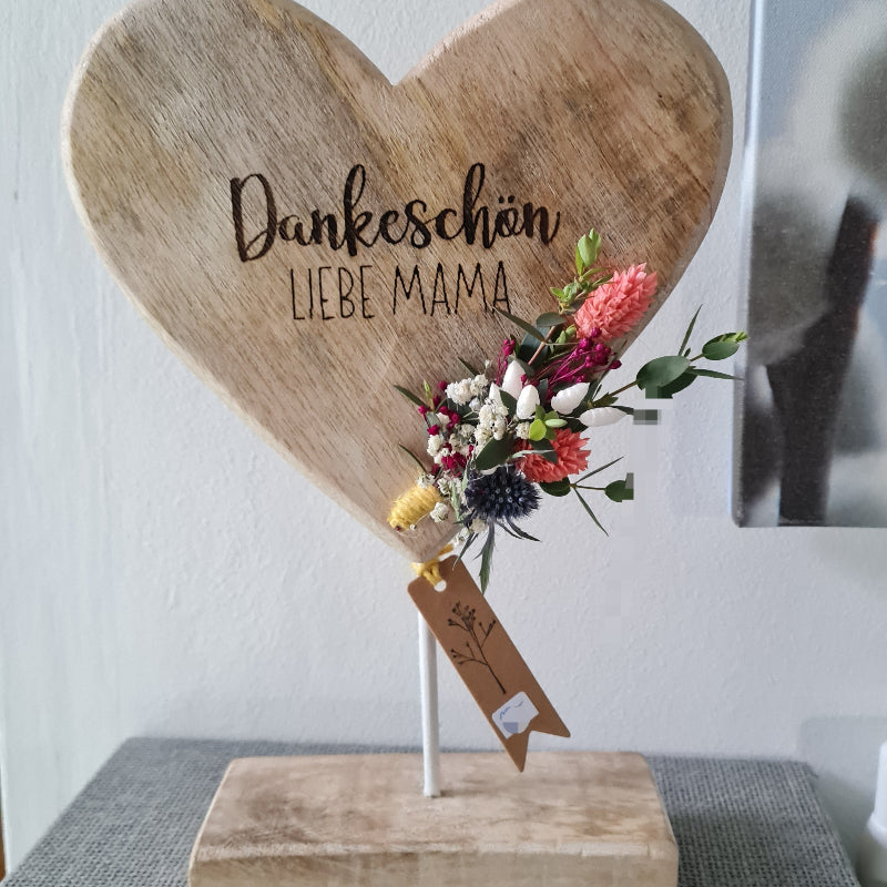 Ein kleines herz aus Holz steht auf einem weißen Metallstab auf einem Holzsockel. Das Herz ist mit persönlichem Wunschtext zum Muttertag graviert und trägt die Aufschrift "Dankeschön liebe Mama". Es wird nach deinem Wunsch gefertigt und mit einem kleinen Strauß haltbarer bunter Trockenblumen dekoriert. Ein beliebtes Geschenk zum Muttertag oder Geburtstag Deiner Mama.