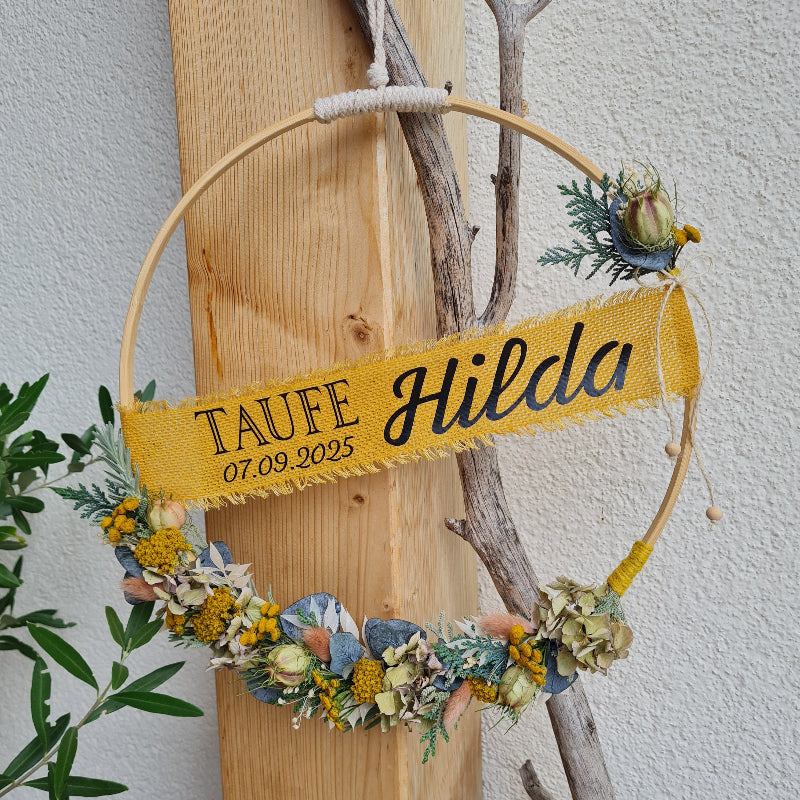 Holzring unten mit Blumen gebunden und mittig ein Spruchband zur Taufe. Der Kranz ist passend zur Jareszeit handgebunden und trocknet schön ein, um eine bleibende Erinnerung zu schaffen. Eine besondere Geschenkidee zu Taufe, Kommunion oder Geburt.