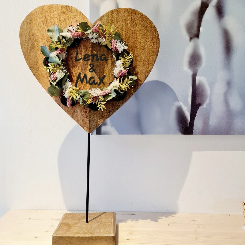 Ein 60 cm großes, massives Holzherz auf Metallständer aus Hochzeitsgeschenk. Wunschtext, Namen und Datum werden personalisiert gelasert. Ein Kranz aus Blumen setzt die Wunschgravur in Szene. Eine beliebte Deko am Geschenketisch.