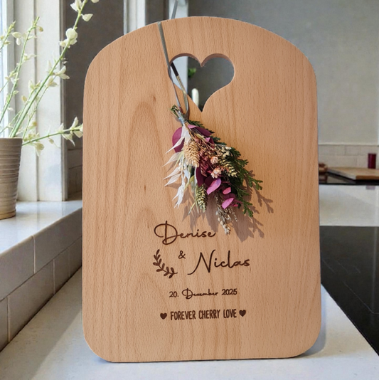 Personalisiertes Holzschneidebrett aus Massivholz mit Gravur der Namen des Brautpaares "Denise und Niklas" sowie dem Datum „Dezember 2025“. Das hochwertige Brett hat eine herzförmige Aussparung oben, ist geschmackvoll dekoriert mit Trockenblumen und eignet sich ideal als Hochzeits- oder Geschenkidee.