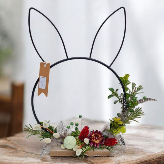 Ein Hasenkopf aus schwarzem Metalldraht, mit Blumen und Grün zum Kranz gebunden steht auf einem Sockel. Eine moderne Hasenfigur für Fensterbrett, Kamin, Konsole oder Ostertisch.