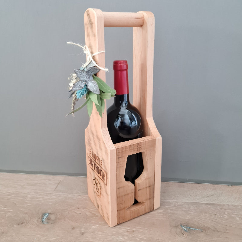 Ein kleiner Träger aus Holz mit Ausschnitt in Form eines Weinglases enthält eine Flasche Rotwein als Geschenk zum Geburtstag. Seitlich ist die Weinkiste mit persönlichem Text graviert "Happy Birthday" und Namen. Das Weingeschenk wird mit einem keinen Anhänger aus Trockenblumen geschmückt.