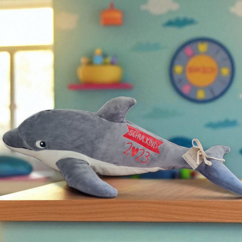 Ein samtweicher Plüschdelfin mit treuherzigem Blick liegt auf einem Schrank im Kinderzimmer eines Schulkindes. Er ist personalisiert mit moderner Schrift und ein ganz besonderes Geschenk zum Schulbeginn.