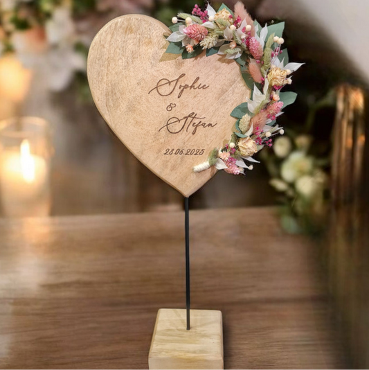 Personalisiertes Holzherz auf einem Standfuß, liebevoll verziert mit einem detailreichen Trockenblumenarrangement auf der rechten Seite. Die Gravur zeigt die Namen sowie das Hochzeitsdatum „24.05.2025“. Ideal als langlebige Hochzeitsdekoration oder Geschenk zur Trauung mit natürlichem Flair.