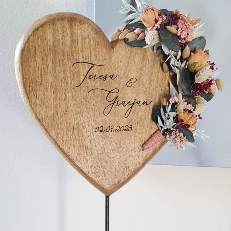 Herz aus Holz auf Metallfuß zur Hochzeit. Das Herz ist personalisiert mit Namen und Datum und liebevoll mit Blumen geschmückt.. Der Name des Brautpaars und das Datum sind in das Holzherz graviert. Ein Kranz aus Trockenblumen umrahmt das Herz und es steht in einer filigran gesteckten Blumenwiese. Ein ganz besonderes Geschenk zur Hochzeit.