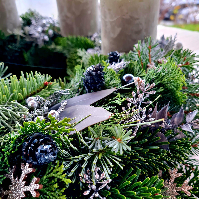 Moderner Adventskranz mit silbernen Details und Tannengrün in Detailansicht. Zoom auf gemischte frische Zweige aus Tanne, Zypresse, Thuje und gefrostete Zapfen und versilberte Frückte von Eukalypzus und Thuje.