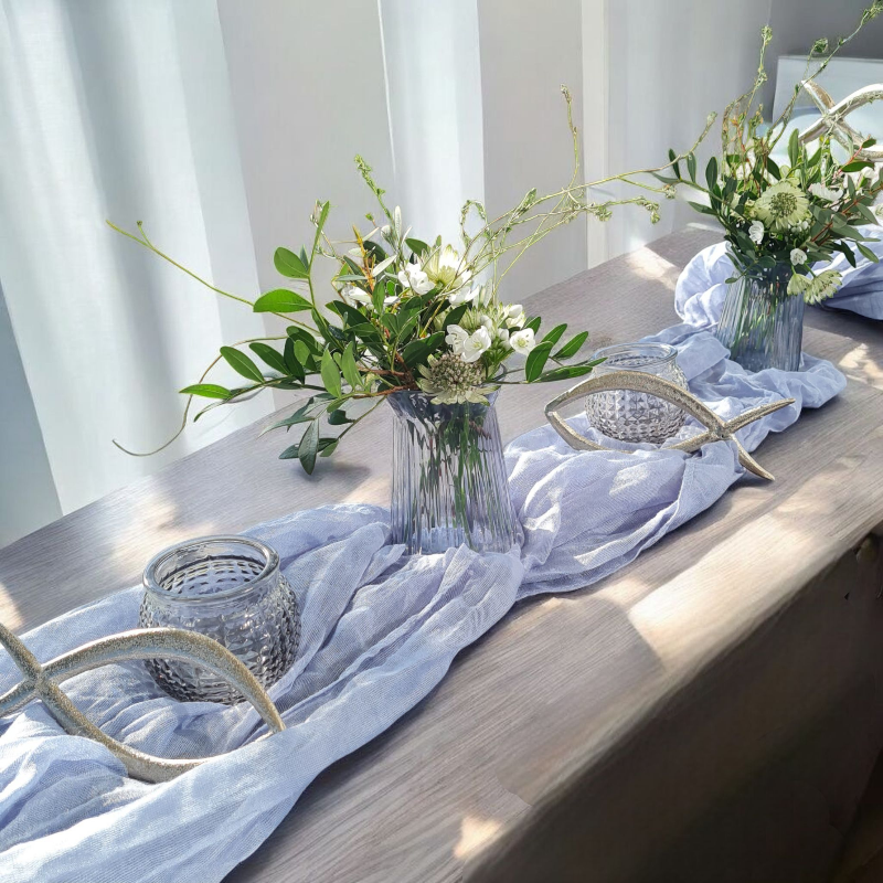 blaue Vasen mit zarten Blüten aus Allium und Sterndolde mit Bättern und Wildkräutern auf blauem Tischband. Dekorierter Tisch für eine Taufe oder Kommunion im angeagten Wildflower Stil. Ein Fischmotiv rundet diese moderne Tischdeko ab. Auch bei Mädchen sehr beliebt.