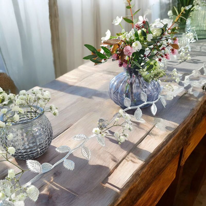 romatische Tischdekoration für eine Hochzeit. Ein zartes Blätterband windet sich um moderne rauchblaue Zwiebelvasen, welche mit Kirschblüten, zarten Blumen und filigranem Gruen gefuellt sind. Sehr modern und romatisch.