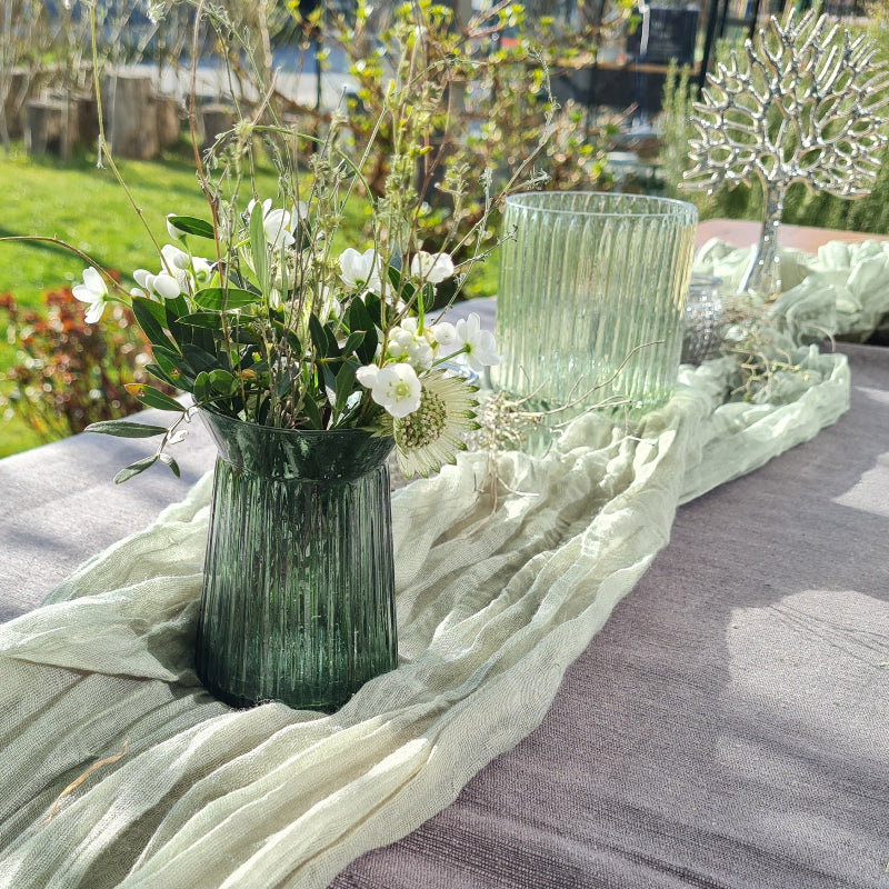 Eine bezaibernde Tischdekoration mit waldgruenen modern geriffelten Glasvasen und Windlichtern. Sehr modern mit salbeigruenem Tischband. Der Baum ist das tragende Symbol dieser Dekoration. Die vasen sind mit zarten Blüten, Blättern und Ästen gefüllt. 